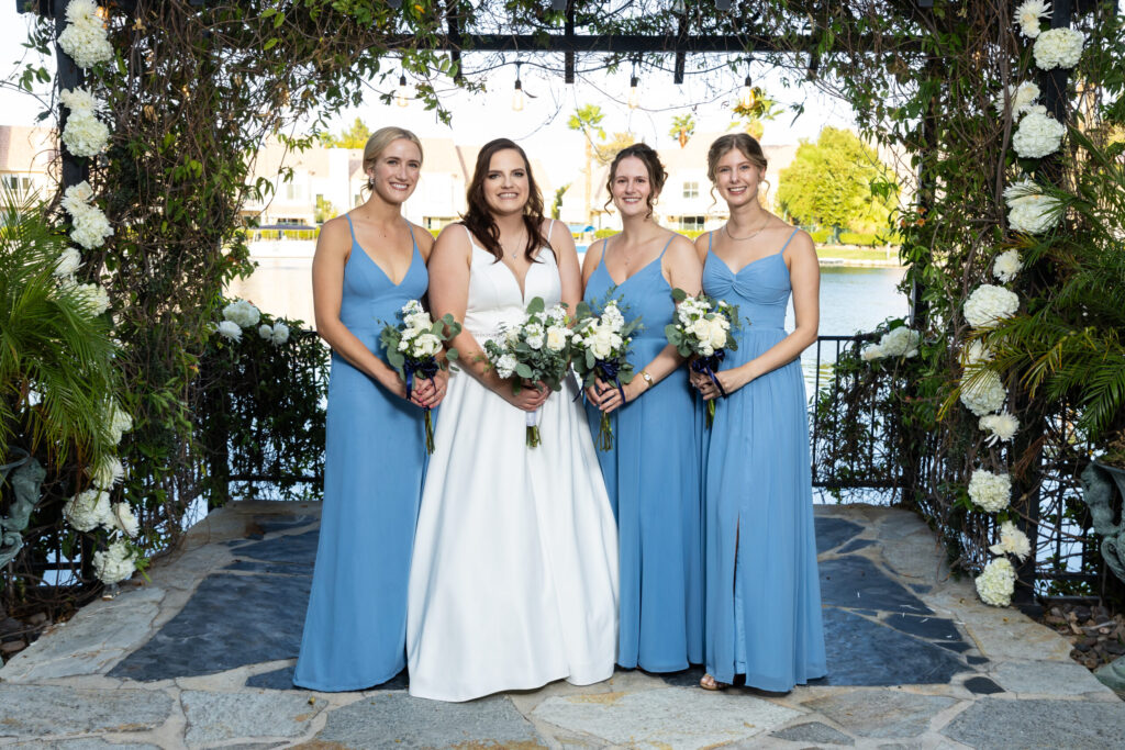 Bridesmaid group posing in blue dresses with bride in white gown, floral backdrop, lakeside setting, capturing wedding celebration essence.