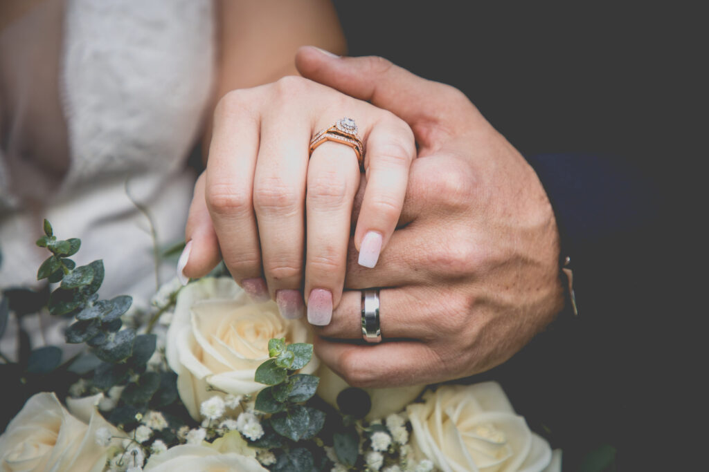 Couple's hands intertwined, showcasing wedding rings and elegant engagement ring, surrounded by white roses and greenery, symbolizing love and commitment for evening weddings in Las Vegas.
